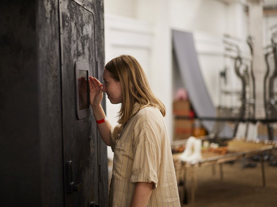A woman in a cream dress looks closely at a display panel in a dark wood panel